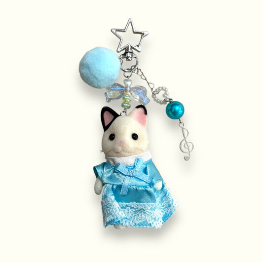 Handmade keychain with Sylvanian Families cat in blue lace dress, pompom, star clasp, bow bead, and music note charm on cream background