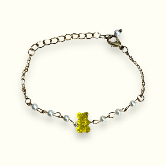 Delicate bracelet with yellow gummy bear charm and pearl accents on a gold chain, displayed on a cream background.