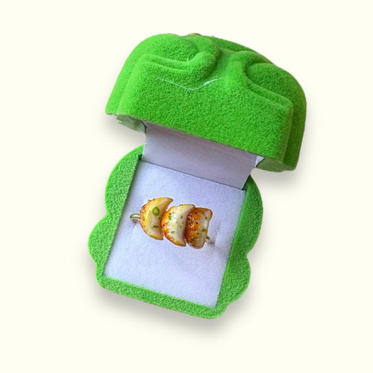 Adjustable ring with three glossy pan-fried dumpling charms and green scallion accents, displayed in a green jewelry box.